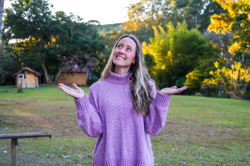 Mari sorrindo e olhando para o céu, com as mãos abertas, em um campo ao entardecer