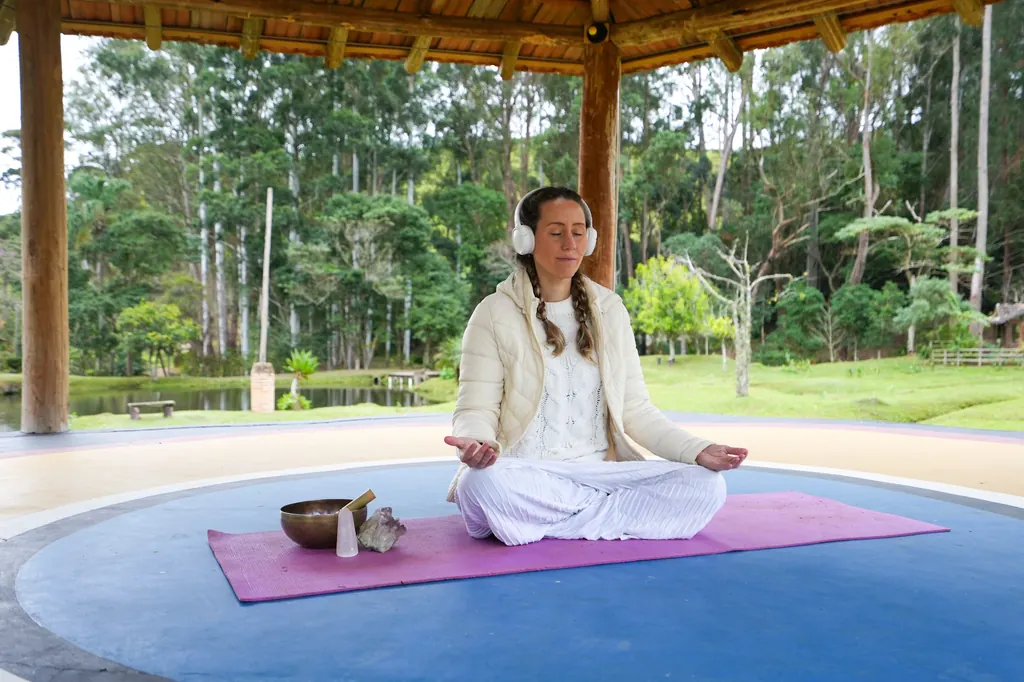 Mari meditando com fones em um quiosque de madeira, com tigela tibetana e cristais ao lado